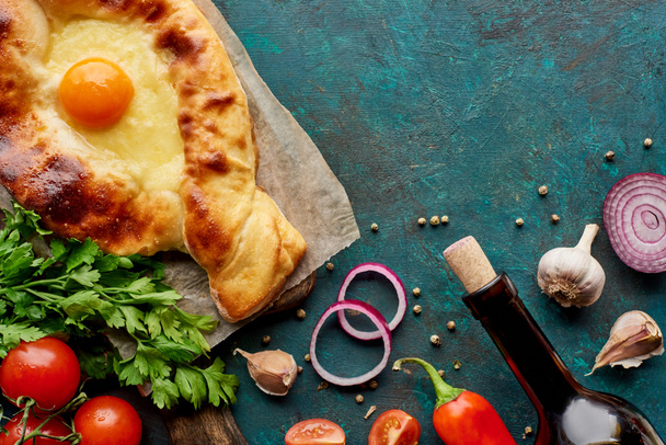 Top view of adjarian khachapuri with wine bottle and vegetables on textured green background - 写真・画像