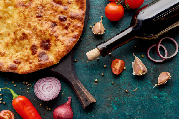 Top view of imereti khachapuri with wine bottle and vegetables on textured green background - Photo, Image