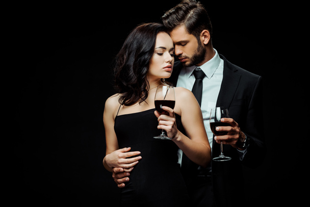 όμορφος άνδρας και ελκυστική γυναίκα κρατώντας ποτήρια με κόκκινο κρασί που απομονώνονται σε μαύρο  - Φωτογραφία, εικόνα