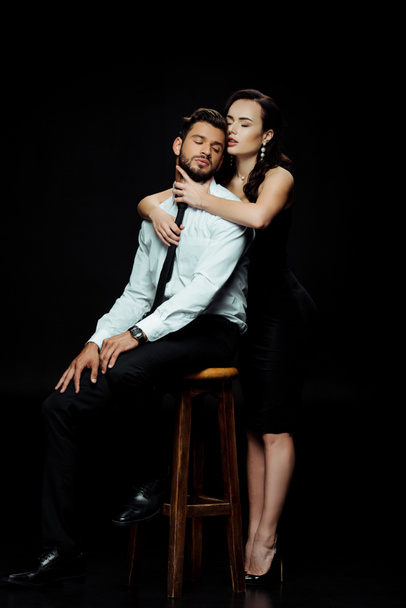 beautiful woman hugging bearded man sitting on chair isolated on black  - Foto, Imagem