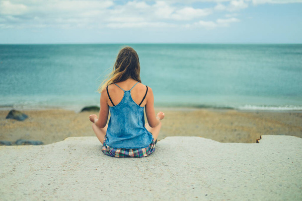 A young woman is sitting in a meditation pose on a concrete pier by the sea - Photo, image