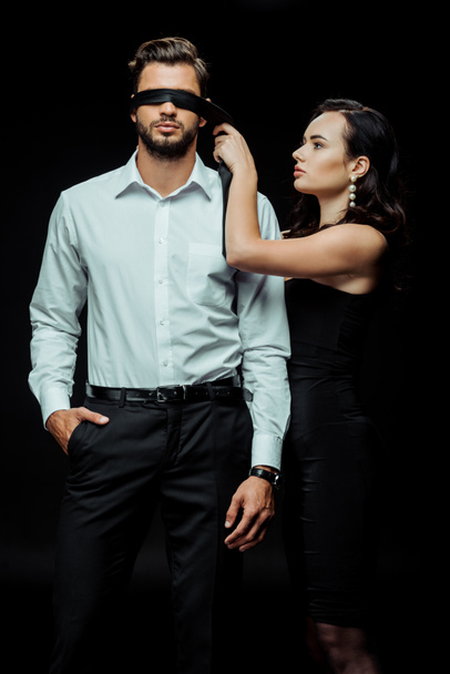 attractive woman wearing blindfold on man standing with hand in pocket isolated on black  - Fotoğraf, Görsel