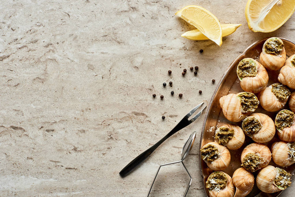 top view of delicious cooked escargots with lemon slices, black peppercorn and tweezers on stone background - Photo, Image