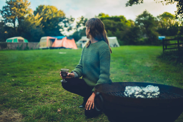 A young woman is enjoying a glass of wine by a fire pit on a camp site on a summer evening - Photo, Image