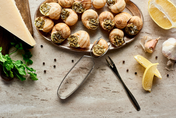 top view of delicious cooked escargots with lemon slices, parsley, black peppercorn and tweezers on stone background - Photo, Image