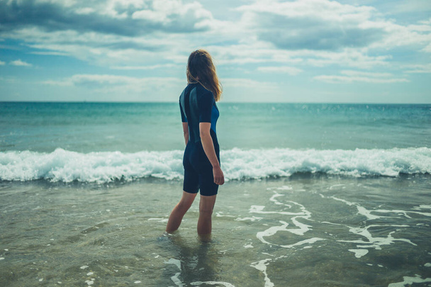 Een jonge vrouw in een wetsuit staat 's zomers op het strand met de golven in - Foto, afbeelding
