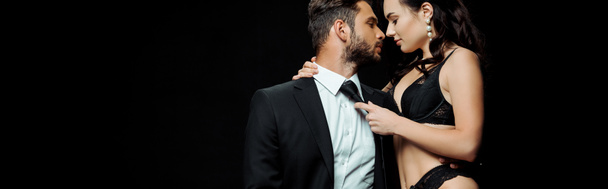 tiro panorámico de mujer en lencería sexy tocando corbata de novio barbudo aislado en negro
  - Foto, Imagen