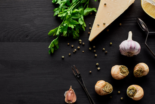 Draufsicht auf köstlich gekochte Escargots auf schwarzem Holztisch mit Zutaten - Foto, Bild