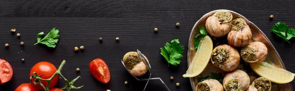 Draufsicht auf köstlich gekochte Escargots auf schwarzem Holztisch mit Zutaten und Pinzette, Panoramaaufnahme - Foto, Bild