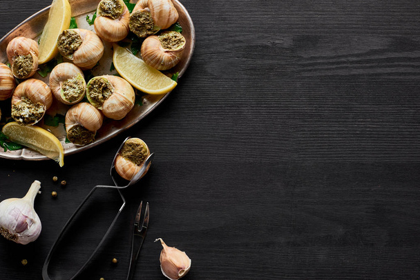top view of delicious cooked escargots with lemon and tweezers on black wooden table - Photo, Image