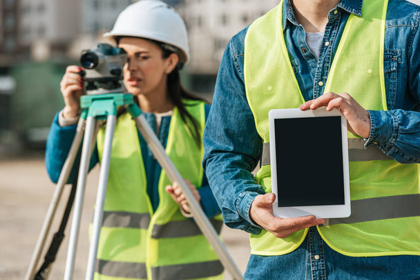 Cropped view of surveyor holding digital tablet with blank screen and colleague with measuring level at background - Photo, Image