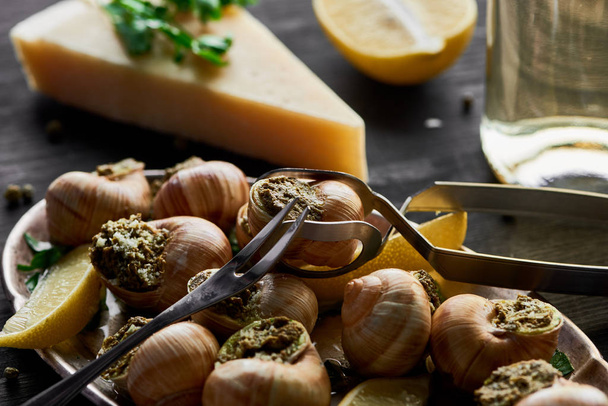selective focus of delicious cooked escargots with lemon, parmesan, cutlery, parsley and white wine on black wooden table - Foto, afbeelding
