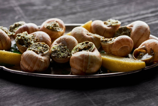 close up view of delicious cooked escargots with lemon slices on plate on black wooden table - Photo, Image
