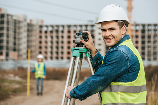Selective focus of smiling surveyor with digital level and colleague at background - Photo, Image