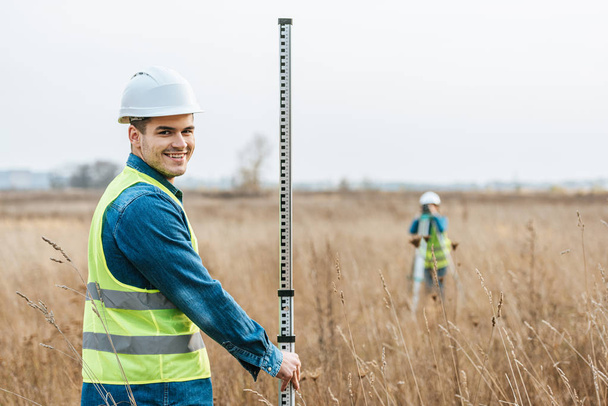 Selective focus of smiling surveyor with ruler and colleague with digital level in field - Photo, Image