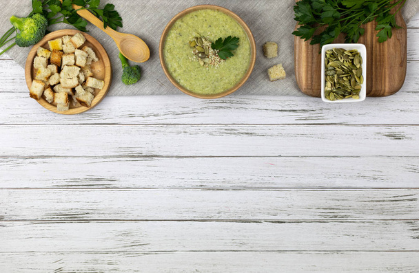 Vegetarische Suppe aus Brokkoli, Kartoffeln, Zwiebeln, Knoblauch, Sesam und Olivenöl sowie Kokosmilch, serviert in einer hölzernen Schüssel. flachlegen eines weißen gealterten Hintergrundes mit hellem Textil - Foto, Bild