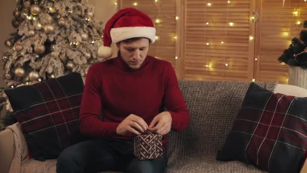 Young Caucasian Man Wearing a Santas Hat sitting on a Sofa near Christmas Tree at Living Room Ties a Bow on a Present Box than Looks to Camera and Smiles. Concept of Holidays and New Year. - Video