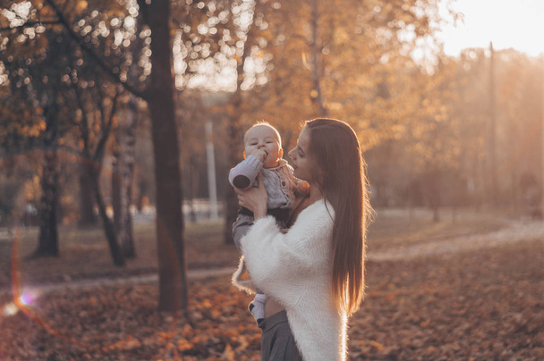 Ευτυχισμένη οικογένεια σε φθινοπωρινή βόλτα. Η μητέρα κρατά τον μικρό γιο στην αγκαλιά της και απολαμβάνει την όμορφη φθινοπωρινή φύση σε ένα πάρκο..  - Φωτογραφία, εικόνα