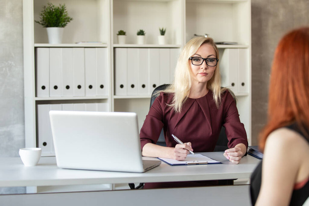 Blond woman in glasses sits behind office table and listen other woman. Selective focus - Foto, imagen