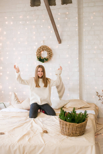 Cute teenage girl has fun at home in a light loft on the bed, decorated for Christmas with garlands and needles. Christmas mood - Photo, Image