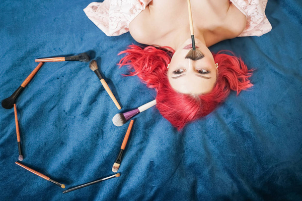 pretty woman with red hair lying with make up brushes and dreaming, top view girl make up master on blue background - Photo, Image