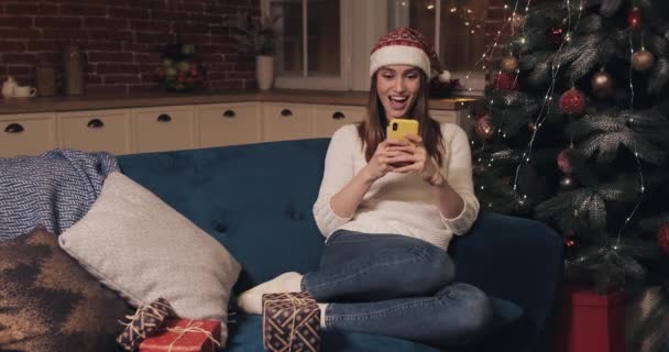 Young Attractive Girl Wearing Christmas Hat Sitting on the Sofa at Home Background Using her Smartphone Rejoices Smiles and Laughs Looking in her Phone Saying Yes - Séquence, vidéo