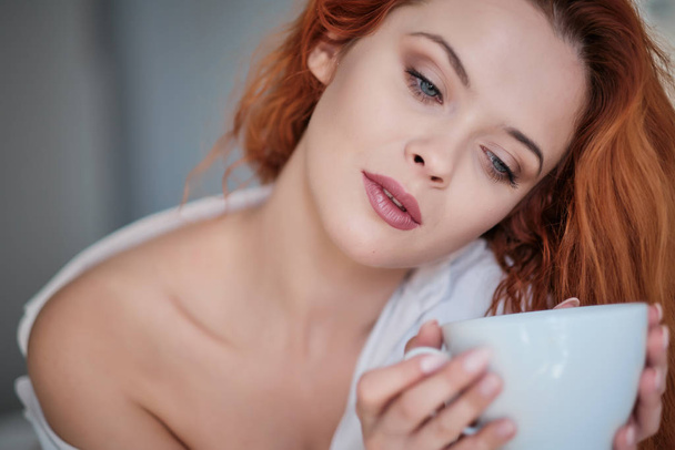 Attractive redhaired woman with a cup of coffee on the bed - Valokuva, kuva