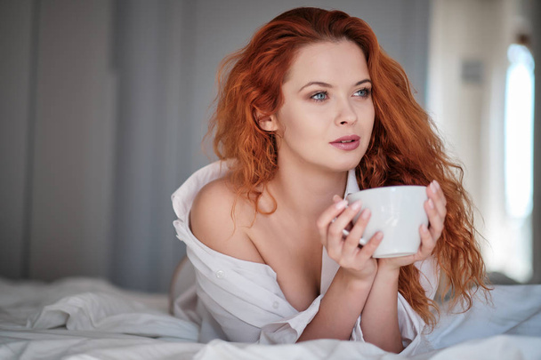 Attractive redhaired woman with a cup of coffee on the bed - Foto, immagini