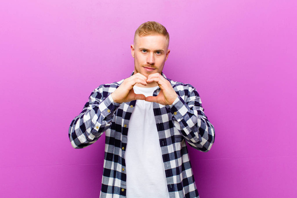 young blonde man with squared shirt smiling and feeling happy, cute, romantic and in love, making heart shape with both hands - Valokuva, kuva