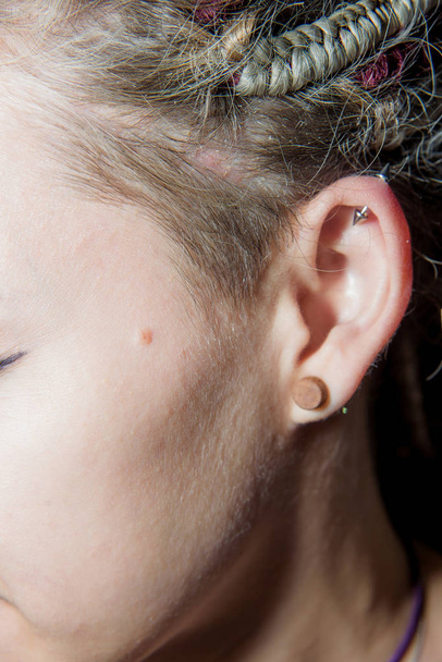Perforación en la oreja de una chica con rastas. Oreja femenina con los pendientes de cerca. Detalles de la imagen de la chica de la cultura informal
 - Foto, Imagen