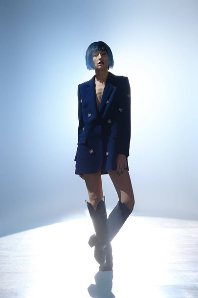 Fashion model with bob haircut in blue jacket with gems decor and in wide high boots on the stage under key light - Фото, изображение