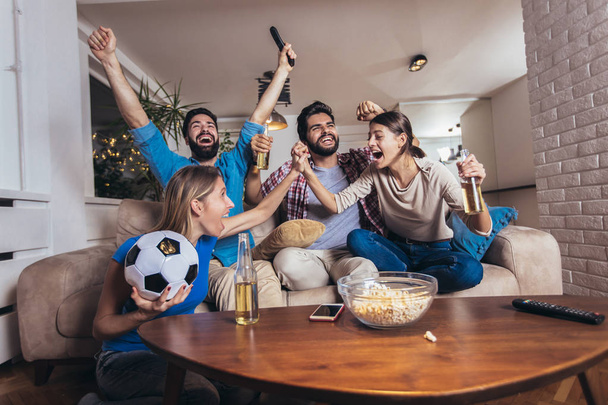 Φίλοι διασκεδάζουν βλέποντας ποδόσφαιρο στο σπίτι, σε εσωτερικούς χώρους. - Φωτογραφία, εικόνα