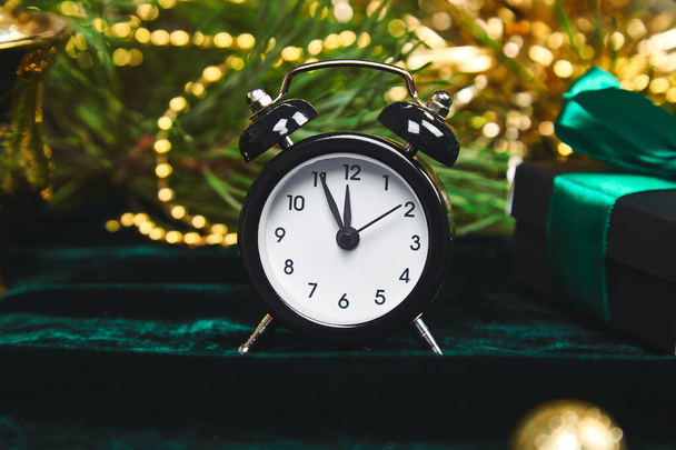 Χριστουγεννιάτικο κουτί δώρου, ξυπνητήρι και κλαδί ελάτης στο τραπέζι. Πάνω άποψη με αντίγραφο χώρο το Νέο Έτος έρχεται. Μαύρο ρολόι και χρυσά χριστουγεννιάτικα στολίδια πράσινο βελούδα, λάμψη φόντο. Έννοια χρόνου Εύα - Φωτογραφία, εικόνα