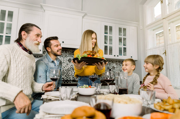 The family sits down for dinner on Thanksgiving. Young woman serves a festive turkey with a salad, grandfather, father and children sit and look at the tasty food and smile - Photo, Image