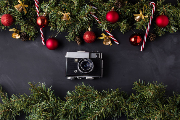 Χριστουγεννιάτικη γιρλάντα κωνοφόρων διακοσμημένη με κόκκινες μπάλες και χρυσά κουδούνια με καραμελένια μπαστούνια στο πάνω και κάτω μέρος του μαύρου τραπεζιού, καθώς και μια vintage κάμερα στο κέντρο. - Φωτογραφία, εικόνα