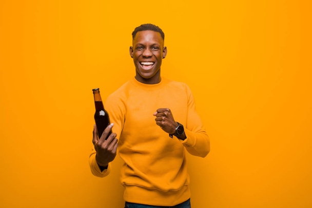 νεαρός Αφροαμερικάνος μαύρος ενάντια στον πορτοκαλί τοίχο πίνοντας μπύρα - Φωτογραφία, εικόνα