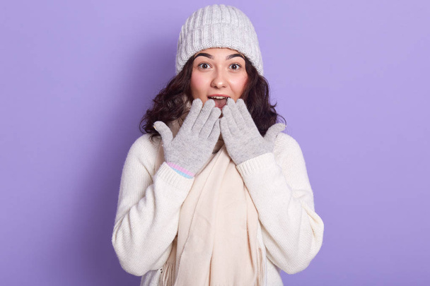 Young attractive girl wearing white sweater, warm cap, scarf and gloves, having light makeup, covering her mouth with her hands, looking with big eyes in surprise, isolated over lilac background. - Foto, immagini