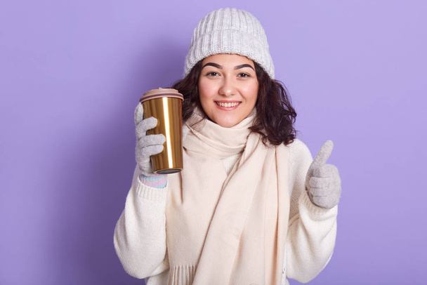 Young woman wearing pale pink scarf, cap, gloves and warm white sweater drinking hot tea or coffee from thermo mug, developing zero waste programs, showing thumb up gesture. Winter season concept. - Фото, зображення