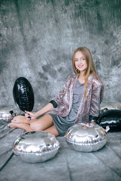 Attractive young girl with blond hair and evening make-up in a silver dress and metallized silver and black balloons. - Фото, изображение