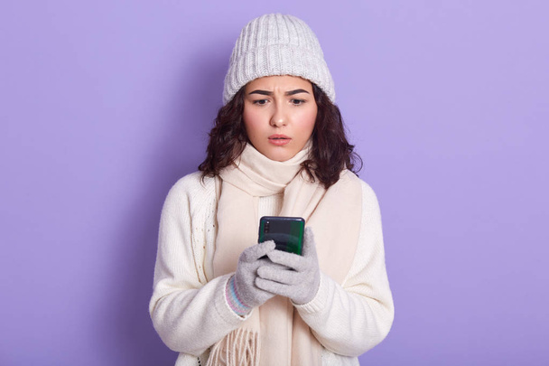 Adorable young girl looking concerned with text message on her smart phone, reads about bad news, has astonished facial expression, wears warm sweater, cap, scarf and gloves, isolated over lilac wall. - Foto, Imagen