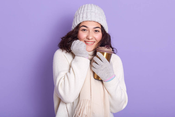 Young attarctive woman basking her hands via cup of hot beverage and gloves, wearing sweater, scarf and cap, posing isolated over purple background, using thermo mug, moving towards zero waste. - Foto, Imagem