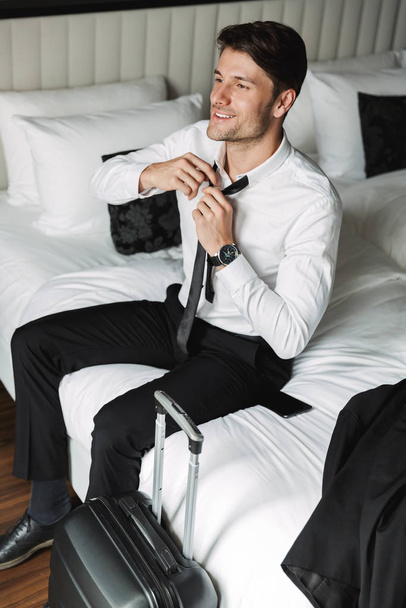 Image of joyful young man unleashing his tie while sitting on be - Fotoğraf, Görsel