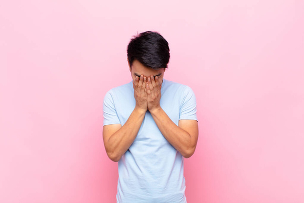 young chinese man feeling sad, frustrated, nervous and depressed, covering face with both hands, crying against flat color wall - Foto, immagini