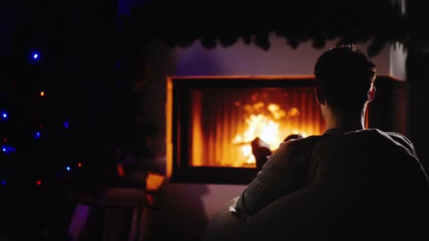 Νεαρός άνδρας με αυτοπεποίθηση ξεκουράζεται δίπλα στο τζάκι και το χριστουγεννιάτικο δέντρο - Πλάνα, βίντεο