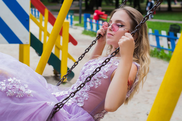 Portrait of young gil wearing dress and violet sunglasses sitting on children swing. tattoes over her arms and face. Close up. City walk. Subculture attributes. - Photo, Image