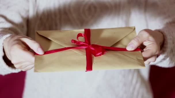 Beautiful girl hands hold a gift envelope with a red ribbon. Beautiful girl holds out a gift envelope with a red ribbon. Gift for the holiday. Happy New Year, Merry Christmas, Happy Valentines Day. - Filmagem, Vídeo