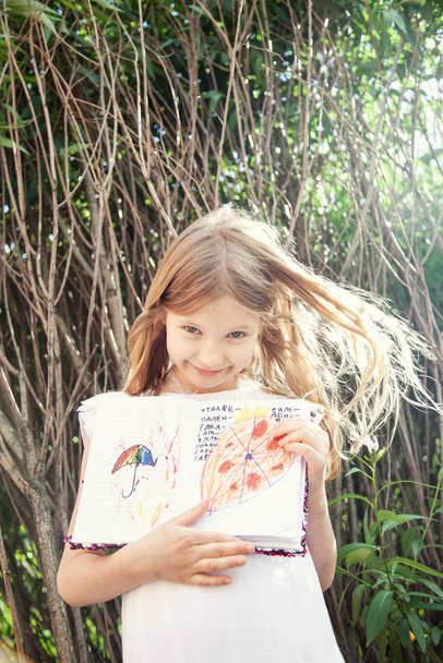 portrait of smiling girl showing drawings near trees in garden - Fotó, kép