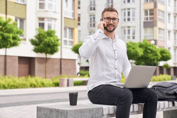Freelancer with laptop sitting on bench talking by phone. - Foto, imagen