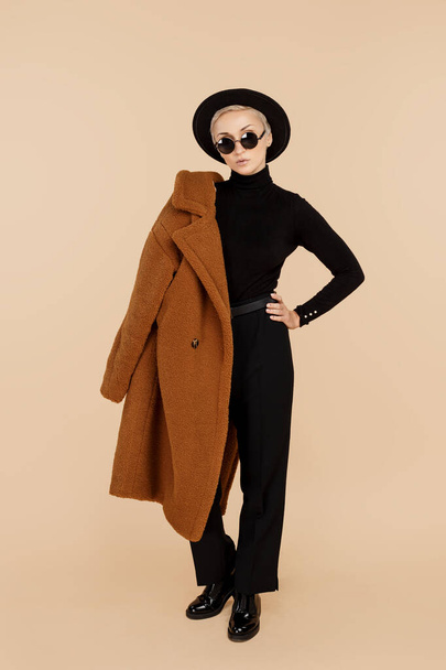 Stylish young hipster blonde woman wearing a coat, hat and sunglasses posing over beige background. Copy space. - Photo, image