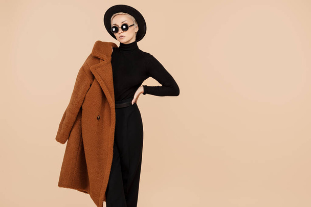 Trendy young hipster blonde woman wearing a coat, hat and sunglasses posing over beige background. Copy space. - 写真・画像